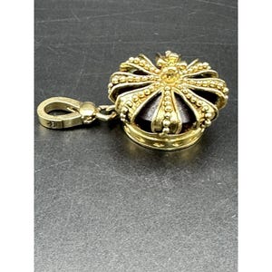 Puede incluir: Un dije de corona dorada con un centro oscuro y un cierre. La corona está detallada con pequeños adornos redondos y una pequeña cruz en la parte superior. El cierre está unido a la corona con una pequeña cadena.