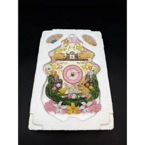 May include: A decorative cuckoo clock featuring a pink clock face and a floral design. The clock is adorned with bunnies, flowers, and a bird, with pink and yellow accents. The clock is in a white foam package.