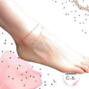 Op de afbeelding: Een delicate, roségouden enkelband met dubbele laag. De bovenste laag heeft kleine kralen, terwijl de onderste laag een kleine hartvormige bedel heeft. De enkelband wordt op een voet gedragen.