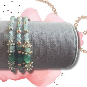 Peut inclure: Trois bracelets en perles avec des perles de pierres précieuses vertes et bleues. Les bracelets sont empilés sur un présentoir en velours gris.