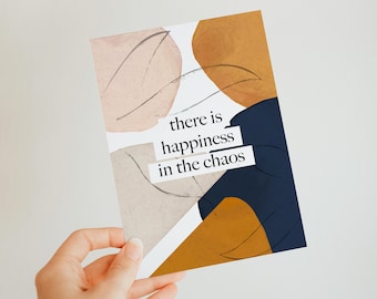 May include: A white card with abstract shapes in shades of brown, beige, and blue. The card has the text "there is happiness in the chaos" printed on it.