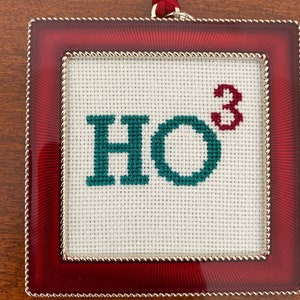 May include: Red framed ornament with a white background and green embroidery that reads "HO" with a superscript "3".