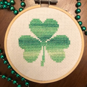 Puede incluir: Un diseño de trébol verde en punto de cruz en un aro de bordado redondo de madera. El trébol es un símbolo tradicional irlandés de buena suerte.