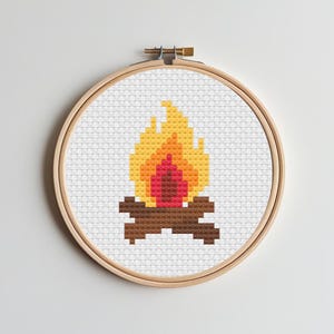 May include: A cross-stitch embroidery hoop with a pixelated campfire design. The flames are in yellow, orange, and red, above brown logs. The artwork is set against a white background.