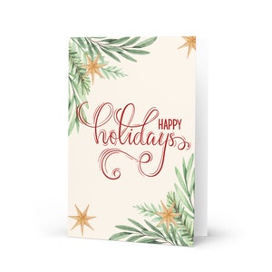 May include: A cream-colored card with a red cursive inscription that reads "Happy Holidays". The card is decorated with green watercolor leaves and gold stars.
