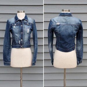 May include: A cropped denim jacket in a medium wash blue, displayed on a mannequin. The jacket features a classic collar, button-up front, and distressed detailing. The back view shows a similar design with a distressed patch.