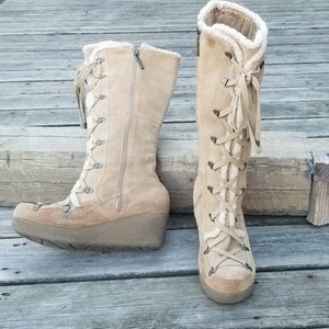 May include: Two tan suede boots with a wedge heel are displayed on a wooden surface. The boots have a lace-up design, a zipper, and a faux fur lining. The boots are designed for cold weather.