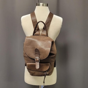 Puede incluir: Una mochila de cuero marrón con cierre de solapa y un pequeño bolsillo frontal. La mochila tiene correas ajustables para los hombros y un asa superior. La bolsa se exhibe en un maniquí.