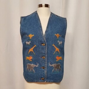 May include: A blue denim vest with a V-neck and button closure. The vest features embroidered images of giraffes, lions, zebras, elephants, and kangaroos. The vest is displayed on a mannequin.