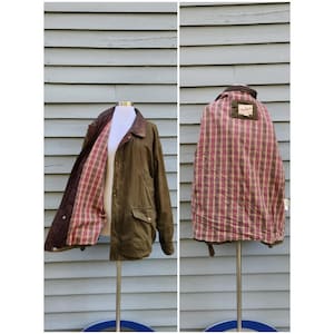 May include: An olive green jacket with a dark brown corduroy collar and a plaid interior lining featuring shades of red, yellow, and green. The jacket includes front flap pockets, a front zipper closure, and a Woolrich brand label inside.