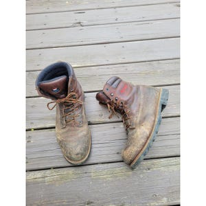 May include: Two brown leather work boots with laces, on a wooden deck. The boots show signs of wear, with scuffs and dirt. The boots have a dark brown collar and a black sole. The boots are made of leather.