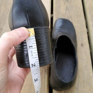May include: Black leather shoe with a block heel. The heel measures approximately 2 inches. The shoe has a rounded toe and a slip-on design. The shoe is shown on a wooden surface.