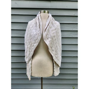 May include: A cream-colored knit shawl featuring an intricate openwork leaf pattern throughout the fabric. The garment is draped over a mannequin, showcasing the detailed texture of the yarn and the symmetrical design of the knitted stitch work.