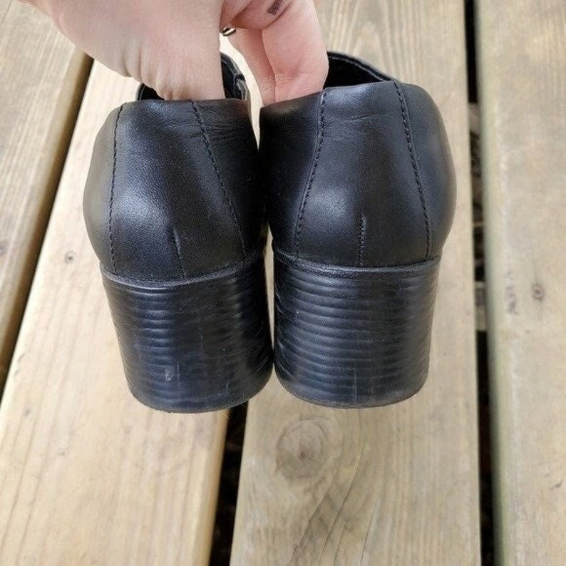 May include: A pair of black leather shoes with a block heel. The shoes have a rounded toe and a strap across the top. The heels are approximately 2 inches high. The shoes are sitting on a wooden surface.