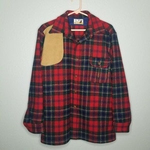 May include: Red, black, and navy plaid flannel shirt with a tan shoulder patch. The shirt has a button-down front, a chest pocket, and a collar. The shirt is hanging on a pink hanger.