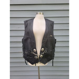 May include: Black leather vest with a V-neck design, featuring two front pockets and decorative silver-toned buttons with a chain. The vest has side laces and a small emblem on the chest. The vest is displayed on a mannequin.