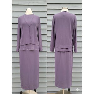 May include: A two-piece lavender suit displayed on a mannequin. The set includes a long-sleeved top with a layered design and a matching long skirt. The top has a V-neckline and a single button closure. The skirt is a straight, floor-length style.