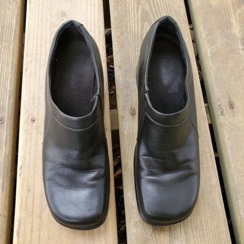 May include: A pair of black leather slip-on shoes with a square toe. The shoes are placed on a wooden surface. The shoes have a low heel and a simple design, suitable for everyday wear.