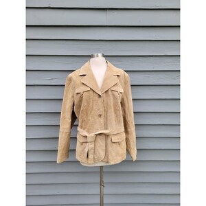 May include: A tan suede jacket with a belted waist and two front pockets. The jacket has a classic collar and button closure. The jacket is displayed on a mannequin against a blue-gray background.