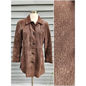 May include: A brown suede jacket featuring a pointed collar, long sleeves, and a front button closure with four dark buttons. The material has a visible textured, matte finish. The jacket is displayed on a mannequin torso against a gray background.