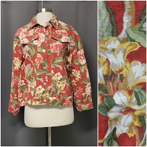 May include: A red denim jacket with a floral pattern featuring pink, white, and yellow flowers and green leaves. The jacket has a collared neckline, button closures, and two chest pockets. The jacket is displayed on a mannequin.