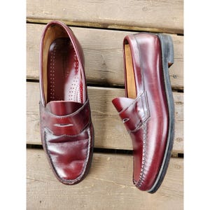 May include: A pair of burgundy leather penny loafers. The shoes have a classic design with a rounded toe, a strap across the vamp, and a low heel. The leather has a glossy finish. The shoes are displayed on a wooden surface.