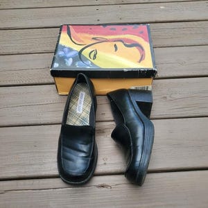 May include: Black leather loafers with a square toe, alongside a shoe box featuring a colourful abstract design. The loafers have a chunky heel and a plaid insole. The shoe box has a yellow and black base.