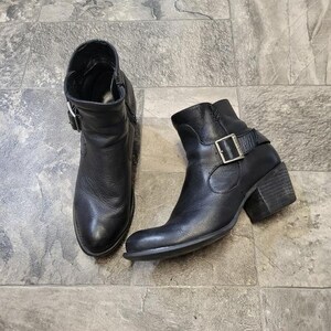 Könnte beinhalten: Ein Paar schwarze Leder-Ankle Boots mit silbernen Schnallen. Die Stiefel haben eine runde Spitze und einen Blockabsatz. Die Stiefel werden auf einer grauen Fliesenoberfläche präsentiert.