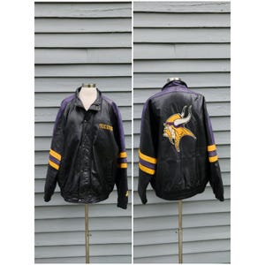 Könnte beinhalten: Schwarze Lederjacke mit lila und goldenen Akzenten. Die Jacke zeigt das Wort "VIKINGS" und ein Wikinger-Logo. Goldene Streifen zieren die Ärmel. Ein Kleidungsstück im Sport-Thema.