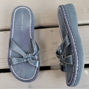 May include: A pair of navy blue sandals with a bow design on the straps. The sandals have a thick sole and white stitching around the edges. The text "LOWER EAST SIDE" is printed on the footbed.