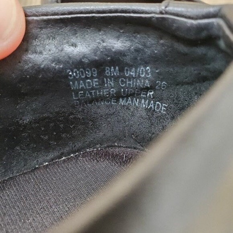 May include: Close-up of the inside of a black shoe. The shoe's interior features stamped text including "30099 8M 04/03", "MADE IN CHINA", "LEATHER UPPER", and "BALANCE MAN MADE". The shoe's interior is dark and textured.