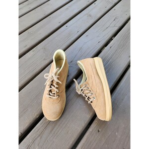 May include: A pair of tan suede sneakers featuring a rounded toe, a lace-up closure with thick tan laces, and a light beige rubber sole. The interior lining of the shoes is a pale cream color, and the exterior is a solid, uniform tan suede material.