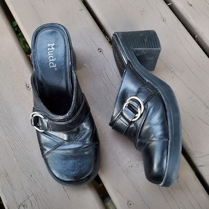 May include: A pair of black leather clogs with a block heel. The clogs have a rounded toe and a strap with a silver buckle. The brand name "Mudd" is visible on the insole. The shoes are on a wooden surface.