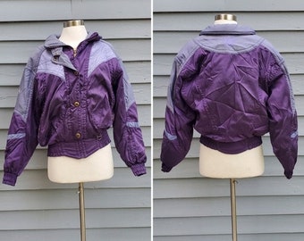 May include: A vintage purple and gray ski jacket with a stand-up collar, button closure, and elastic cuffs. The jacket is displayed on a mannequin and features a cinched waist and a quilted design. The jacket is a retro style.
