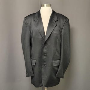 May include: A dark gray blazer with a three-button closure, a notched lapel, and two front pockets. The blazer is displayed on a mannequin. The sleeves have buttons at the cuffs. The fabric has a slight sheen.