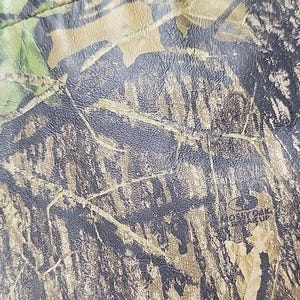 Op de afbeelding: Close-up van een camouflagepatroon met tinten groen, bruin en zwart. Het patroon verschijnt op een getextureerd oppervlak, met de woorden "Mossy Oak Break Up" zichtbaar.