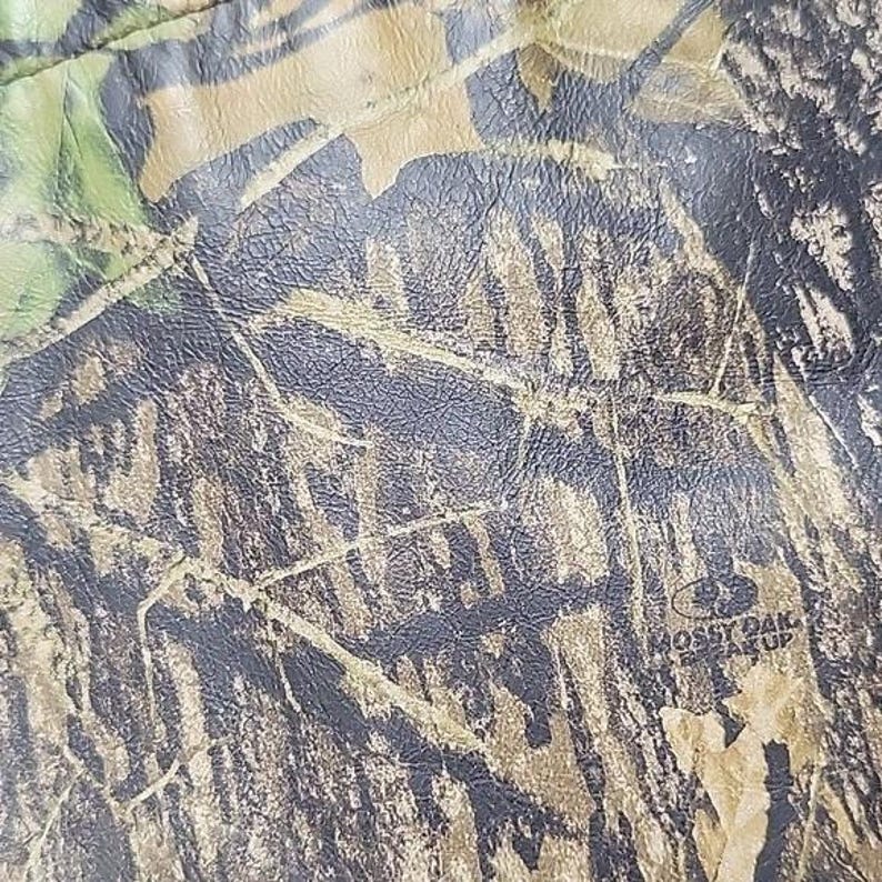 Op de afbeelding: Close-up van een camouflagepatroon met tinten groen, bruin en zwart. Het patroon verschijnt op een getextureerd oppervlak, met de woorden "Mossy Oak Break Up" zichtbaar.