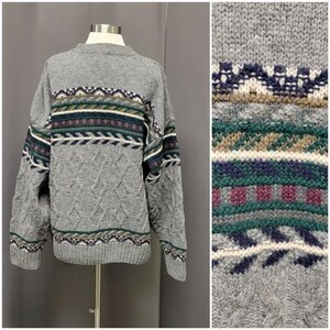 May include: A grey cable-knit jumper with a crew neck and patterned bands across the chest. The bands feature geometric designs in navy, green, burgundy, and cream. The jumper is displayed on a mannequin.