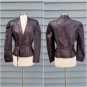 May include: A dark brown leather jacket with a double-breasted front, a belted waist, and puffed sleeves. The jacket is displayed on a mannequin against a blue-gray background. The jacket has a classic, tailored design.