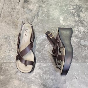 May include: A pair of brown leather sandals with a wedge heel. The sandals have crisscross straps and a buckle closure. The brand name "MIA" is visible on the insole. The wedge heel is approximately 3 inches high.