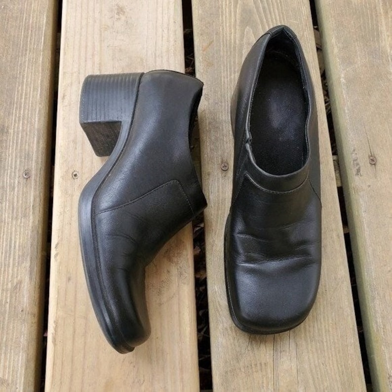 May include: A pair of black leather clogs with a square toe and a block heel. The shoes are displayed on a wooden surface. The clogs have a slip-on design and a rounded vamp.