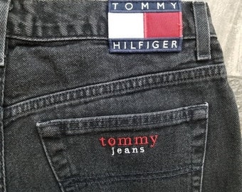 May include: Detail of black denim jeans. The jeans have a small back pocket with "tommy jeans" embroidered in red. A rectangular patch with the "Tommy Hilfiger" logo is sewn on the waistband.