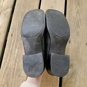 May include: A pair of black Clarks shoes, viewed from the soles up. The shoes have a square toe and a small heel. The Clarks logo is visible on the heel. The shoes are on a wooden surface.