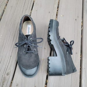 May include: Pair of black suede Guess shoes with a lace-up design and a chunky platform heel. The shoes have a rounded toe and a rubber sole. The Guess logo is visible on the insole.