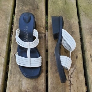 May include: Pair of Fashion Bug Flexi Sole sandals. The sandals have a black sole and a white strap design. The sandals are on a wooden surface.