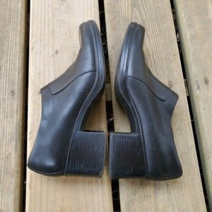 May include: A pair of black leather clogs with a unique design. The shoes have a square toe and a chunky, open heel. The clogs are set on a wooden surface.