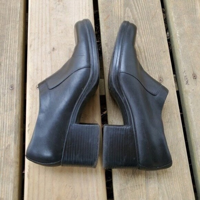 May include: A pair of black leather clogs with a unique design. The shoes have a square toe and a chunky, open heel. The clogs are set on a wooden surface.