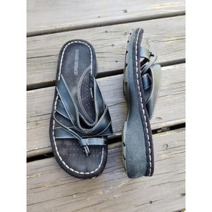 May include: Pair of black sandals with a thong toe post and multiple straps. The sandals feature a textured footbed and a thick sole. The brand name "Rugged Outback" is visible. White stitching details the edges of the sandals.