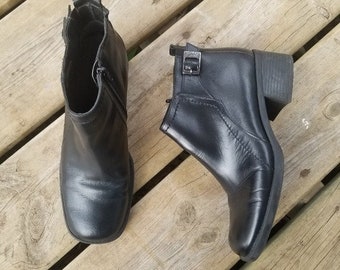 May include: Two black leather ankle boots with square toes and block heels. One boot has a side zipper, and the other has a buckle detail. The boots are set against a wooden background.