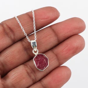 Puede incluir: Un collar de cadena de plata con un colgante con una piedra preciosa de rubí rojo. El rubí está facetado y tiene una forma natural irregular.